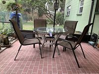 Outdoor patio setup showing round glass top table with white wicker style base and four brown sling chairs with dark metal frames on red brick patio.