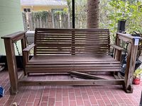 Wooden porch swing showing front view with slatted design and armrests, sitting on brick floor near railing and greenery.