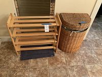 Wooden 4-shelf shoe rack and wicker hamper set on tiled floor. Shoe rack natural wood finish with horizontal slat shelves, hamper is cylindrical wicker with hinged lid and fabric liner visible.