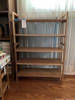Wooden shoe shelf with five slatted shelves against a window with a curtain. Shelf has a natural wood finish and a simple frame structure.