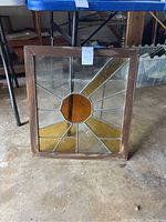 Stained glass window with wooden frame placed on the floor, showing sun ray design with amber central circle and yellow and clear glass panels.
