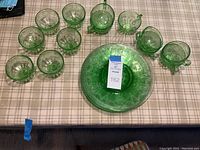 Top-down view of vintage green depression glass cups and one 10-inch plate with floral pattern, one plate has blue tape indicating chip.