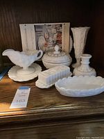 Milk glass items arranged on wooden shelf, showing gravy boat and underplate, vase, covered dish, butter dish, bowl, and candlestick holder.