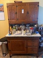 Front view of oak Hoosier cabinet showing upper and lower cabinets with white enamel work surface covered in miscellaneous contents