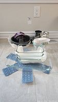 Photo showing black Bundt pan in packaging, metal sifter, two metal mini loaf pans, Pyrex glass baking dishes stacked, and blue silicone trivets on floor.