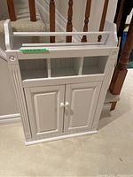 Front view of white wall mount bathroom cabinet showing two doors with knobs, three open top compartments and towel bar.