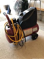 Front angle showing the red tank, motor housing, attached yellow air hose, and wheels of the compressor