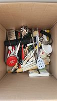 Top down view of box filled with assorted cooking utensils including red Betty Crocker measuring cups, wooden spoons, spatulas, can opener and pot brush.