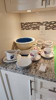 Three nesting mixing bowls with one blue interior, Corelle plates and multiple teacups with saucers on kitchen counter
