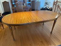 Photo showing full view of the oval solid wood dining table with chairs in background, highlighting tabletop and legs.