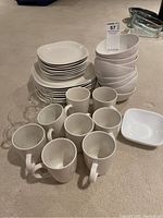 Photo showing multiple white ceramic dinner plates, smaller plates, mugs and bowls arranged on a carpet surface.
