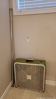 Photo of the Rotor box fan with white front grille and olive green control panel, placed on floor next to metal shower rod in corner against wall.