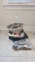 Photo showing stacked metal mesh strainers inside a black roasting pan with some packing papers. A bundle of metal forks, spoons, and knives is in front.