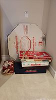 Stack of board games with large octagonal Crokinole board box upright behind them, includes multiple game boxes stacked on carpeted floor.