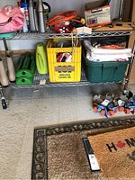 Metal rack with various gardening supplies, including a yellow crate with tools, green watering can, and two green sprinklers under the rack.