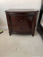 Front view of solid wood night table with two drawers and round knobs in dark brown finish on carpeted floor.