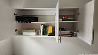 White wall-mounted cabinet open to show shelves with desk organizer, printer papers, books, and software boxes.