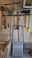 Full view of hanging outdoor tools including shovels, rakes, and the snow scoop in front of a wall with a beige couch nearby.