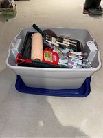 Full view of gray plastic bin containing assorted painting supplies such as brushes, tape measure, painter's tape, paint roller, and paint tubes.