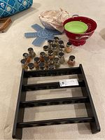Photo showing black wooden spice rack on floor with sixteen small spice jars around it, also blue trivets, covered bowl wrapped in paper, and red colander bowl with green insert visible.