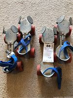 Two pairs of vintage metal frame roller skates with red rubber wheels and blue adjustable straps shown from above on carpeted floor.
