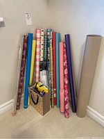 Multiple rolls of wrapping paper standing upright against a wall, various colors and patterns visible with a gift bag at the base containing tissue paper and ribbons.