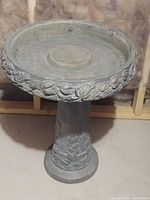 Frontal view of cast concrete bird bath showing round basin and pedestal with leaf and vine decoration and brass tones