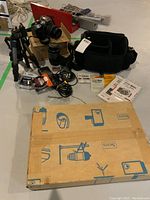 Overview photo displaying all items included in the lot arranged on the floor showing camera, lenses, tripod, carry bag, manuals, charger, memory cards and the slide sorter box.