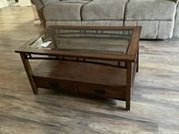 Front view showing the rectangular coffee table with glass top, wood frame, metal trim, and two drawers with metal handles beneath the top glass surface.