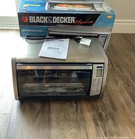 Front view of Black & Decker convection oven with glass door, control panel, power cord, instruction manual, and original box behind it.