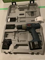 Ryobi cordless drill, battery, charger, and drill bits inside a gray plastic carrying case, showing overall contents of the lot.