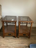 Two matching wood end tables with glass inset tops and metal trim, lower shelf, and drawer positioned side by side on wood flooring.