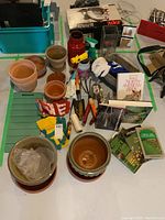 Overview of lot items arranged on table including unglazed clay pots, glazed ceramic planters, garden gloves, gardening tools (trowels, garden claw), several gardening books, a garden kneeling pad, gardening chemicals and spray bottle.