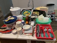 Photo shows stacked and grouped kitchenware items on a table including various bowls, plastic containers, a salad spinner, a clock, and silverware in a red organizer.