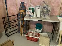 Photo showing ironing board, crutches, quilt rack, T-fal iron, paper towel holder, plastic bin, bath rug, and other items on floor and table in basement setting.