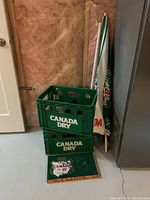 Photo of two green Canada Dry branded plastic pop cases stacked, with partial view of umbrella, mat, and wall.
