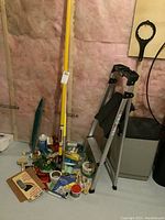 Photo showing step ladder, extension poles, and clustered painting supplies on floor including tapes, brushes, rollers.