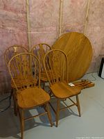 Photo showing four solid wood dining chairs clustered together with the round table top leaning against the wall. Table legs are on the floor beside the chairs.