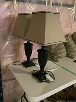 Pair of matching table lamps with black urn-shaped metal bases and trapezoidal beige fabric shades placed on white table in basement setting with pink insulation in background.