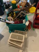 Photo showing green plastic bin filled with assorted tools including hand saws, extension cords, power strip, and gloves; white plastic heat duct covers are in front of the bin