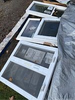 Four white aluminum framed casement windows with attached screens, placed on pallet and ground, some have stickers and labels visible.