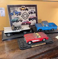 Photo of the three model cars displayed on a wooden surface with the Chevy Trucks metal sign in the background.