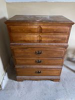 Front view of wooden chest of drawers showing four drawers with missing pulls on top drawers and metal handles on lower drawers. Visible stains and scratches on top surface.