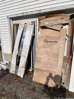 Photo showing multiple storm doors and Showerlite shower door packaging leaning against a building, packaging exposed to elements.