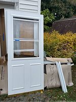 White screen door leaning against a pack of insulation outside, features a glass window and white frame.