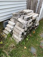 Stack of weathered concrete garden edging stones stacked outdoors along a house wall surrounded by grass.