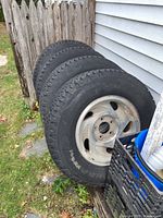 Four Goodyear truck tires with steel rims leaning against house corner outside next to crate and ladder.