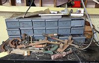 Wide view of the metal storage cabinet with assorted tools placed on the workbench in front. Shows rusty horseshoes, hand tools, hooks, and other hardware items.