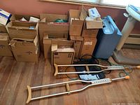 Photo showing multiple cardboard boxes containing bandages, calf supporters, and medical supply packaging, alongside two pairs of aluminum crutches with wooden grips resting on the floor near a blue medical container.