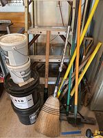 Photo of the Husky wooden 8-foot ladder behind buckets of adhesive, broom, and several garden tools including a shovel and hoe with wooden handles.
